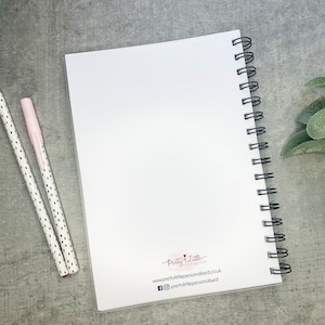 K&ouml;nnte beinhalten: Ein wei&szlig;es Spiralblock mit einem leeren Umschlag. Der Block hat eine schwarze Drahtbindung und ein kleines Logo auf der R&uuml;ckseite, das "Pretty Little" mit einer Website-Adresse und Social-Media-Handles sagt.
