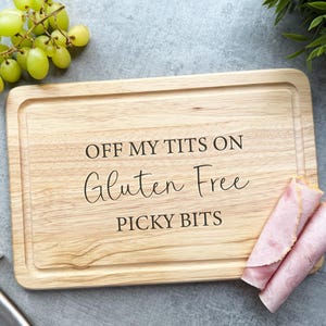 May include: A wooden chopping board with the text "OFF MY TITS ON Gluten Free PICKY BITS" engraved. The board has a groove around the edge. Green grapes and rolled ham are also visible.