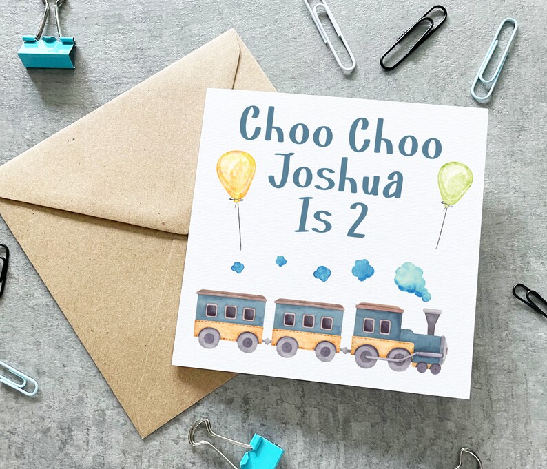 May include: A white card with blue text that reads "Choo Choo Joshua Is 2". The card features a blue and yellow train with yellow and green balloons.