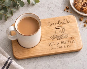 May include: A light-colored wooden serving board with a circular cutout holding a speckled ceramic mug filled with tea. The board is engraved with the text Grandad's TEA & BISCUITS Love Jack & Rosie and a line drawing of a mug and three biscuits.