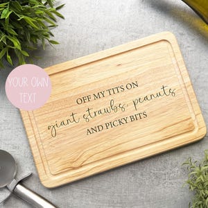 May include: A rectangular wooden cutting board with a groove around the edge. The board has the text "OFF MY TITS ON giant strawbs, peanuts AND PICKY BITS" engraved on it. A pizza cutter is in the lower left corner.