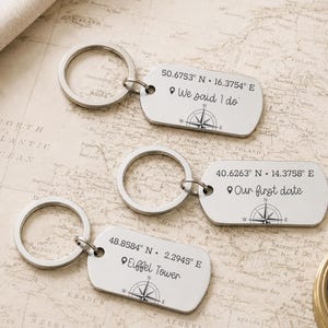 May include: Three silver-toned metal rectangular keychains with rounded corners, each featuring a split ring. The tags are engraved with geographic coordinates, a location pin icon, custom text including "We said I do," "Our first date," and "Eiffel Tower," and a compass rose design.