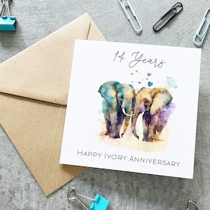 May include: A white card with a watercolor illustration of two elephants standing close together. The elephants are painted in shades of blue, purple, and brown. The card says "14 Years Happy Ivory Anniversary" and has small hearts scattered around the elephants.