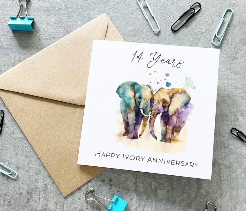 May include: A white card with a watercolor illustration of two elephants standing close together. The elephants are painted in shades of blue, purple, and brown. The card says "14 Years Happy Ivory Anniversary" and has small hearts scattered around the elephants.