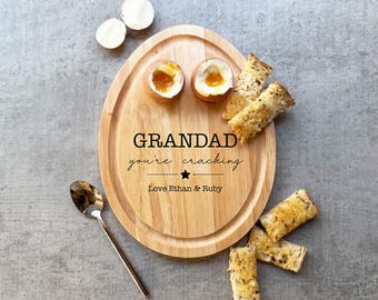 May include: Wooden serving board with the text "GRANDAD you're cracking" and the names "Love Ethan & Ruby". The board holds two halves of a soft-boiled egg and several pieces of toast. A spoon rests on the side.