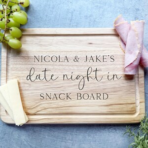 May include: A wooden snack board with the text "NICOLA & JAKE'S date night in SNACK BOARD" in black script. The board is surrounded by grapes, cheese, and rolled deli meat. The board is rectangular with a recessed edge.