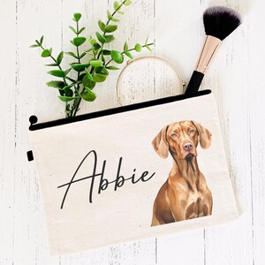 May include: A canvas makeup bag with a black zip and handle. The bag features a watercolour portrait of a dog and the name "Abbie" in script. A makeup brush and greenery are placed next to the bag.