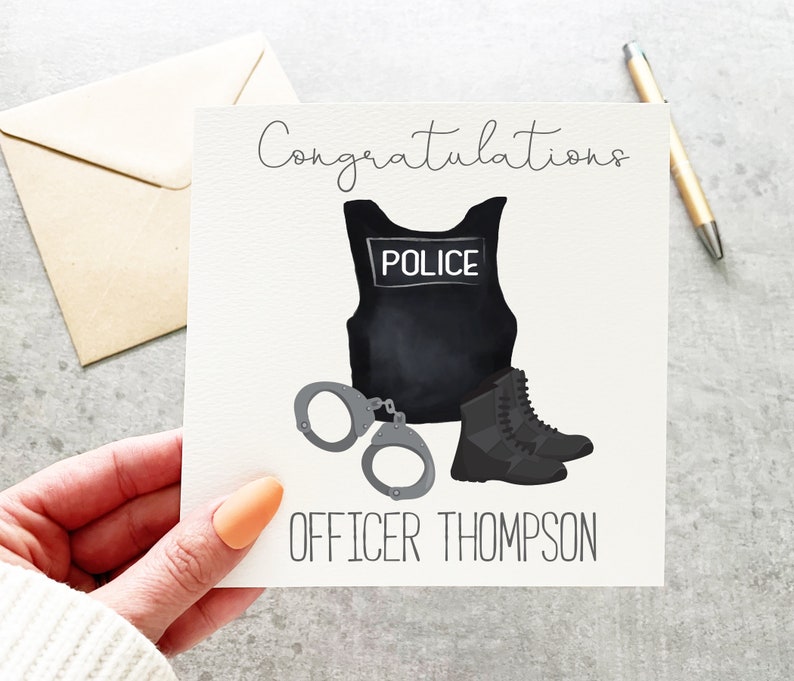 Op de afbeelding: Gefeliciteerd kaart met een zwart-witte illustratie van een politiekostuum, inclusief een kogelvrij vest met het woord "POLICE" erop, handboeien en laarzen. De tekst "OFFICER THOMPSON" staat onder de illustratie.