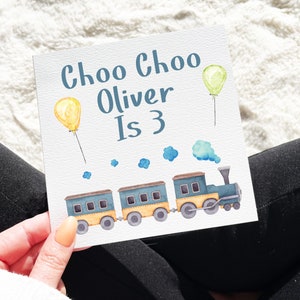 May include: A white card with the text "Choo Choo Oliver Is 3" in blue lettering. A blue and yellow train with three cars and a smoke stack is illustrated on the card. There are yellow and green balloons with strings.