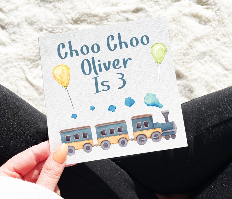 May include: A white card with the text "Choo Choo Oliver Is 3" in blue lettering. A blue and yellow train with three cars and a smoke stack is illustrated on the card. There are yellow and green balloons with strings.