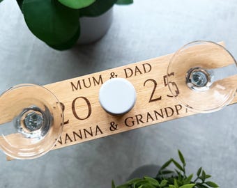 May include: A wooden wine glass holder with the words "MUM & DAD 25 NANNA & GRANDPA" engraved on it. Two wine glasses are in place, and a white bottle stopper sits in the center. The holder is surrounded by green plants.