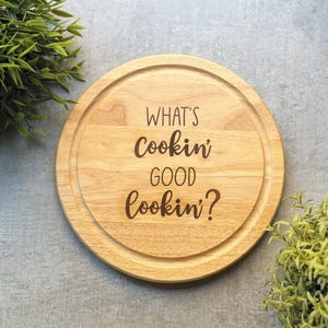 May include: A round wooden cutting board with a groove around the edge. The text "WHAT'S Cookin' GOOD Cookin'?" is etched into the wood. The board is light brown and sits on a gray surface.