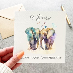 May include: A card with two watercolor elephants standing next to each other with their trunks intertwined. The elephants are painted in shades of blue, purple, and orange. The card says "14 Years Happy Ivory Anniversary".