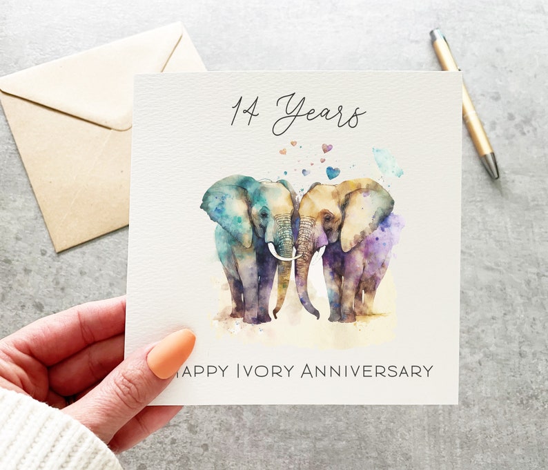 May include: A card with two watercolor elephants standing next to each other with their trunks intertwined. The elephants are painted in shades of blue, purple, and orange. The card says "14 Years Happy Ivory Anniversary".