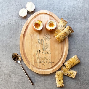 May include: A wooden egg-shaped serving board with the words "Mum's Dippy Eggs" and a daffodil illustration. The board holds two soft-boiled eggs, toast soldiers, and a spoon. The eggs have runny yolks.