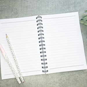 May include: A white spiral bound notebook with lined pages. Two pens, one pink with white polka dots and one black with white polka dots, are lying on the open notebook.