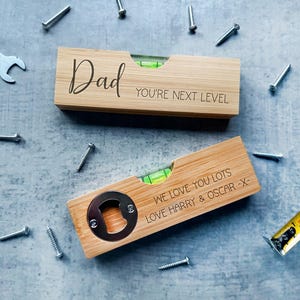May include: Two wooden spirit levels with engraved text. One says "Dad, You're Next Level." The other has a bottle opener and reads "We love you lots, Love Harry & Oscar -X-". Screws, a spanner, and a tape measure are scattered around.
