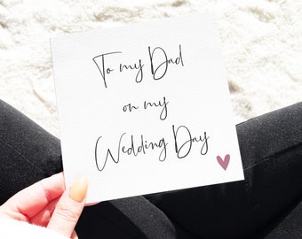 May include: A square white greeting card with black cursive text that reads To my Dad on my Wedding Day, featuring a small dusty rose colored heart icon in the bottom right corner. The card has a textured paper finish and is held in a hand.