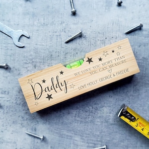 May include: A wooden spirit level engraved with the word "Daddy" and star motifs. The level features a green bubble and the inscription "WE LOVE YOU MORE THAN YOU CAN MEASURE LOVE HOLLY, GEORGE & FREDDIE". A yellow tape measure and screws are scattered around.