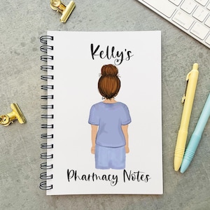 May include: A white spiral bound notebook with a blue and white illustration of a woman with brown hair. The notebook has the text "Kelly's Pharmacy Notes" printed on the front.