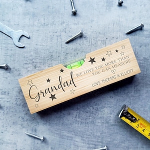 May include: A wooden spirit level engraved with the word "Grandad" and the message "WE LOVE YOU MORE THAN YOU CAN MEASURE". The level is surrounded by tools including screws, a spanner, and a tape measure.
