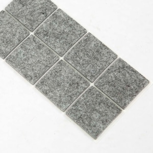 May include: A collection of twelve square, grey felt furniture pads arranged in a grid. The pads have a textured appearance, designed to protect floors from scratches. The pads are on a white background.