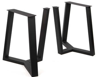 May include: A pair of matte black metal table legs featuring a geometric, angular frame design. Each leg assembly consists of two vertical supports connected to a flat mounting plate at the top and a V-shaped base at the bottom.