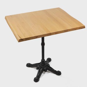 May include: A square, light-coloured wooden table top is supported by a black, ornate, cast-iron base. The table's top is approximately 76 cm wide and 76 cm long. The base has a four-legged design.