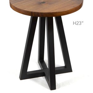 May include: A round side table with a natural brown wood grain top and a black metal base featuring four angled legs connected by a cross-shaped support at the bottom. The text H23" is printed on the right side, indicating a height of 23 inches.