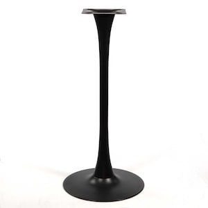 May include: A matte black metal table base featuring a wide circular floor plate, a slender, tapered central column, and a square mounting plate at the top with four pre-drilled holes for securing a tabletop to the structure.