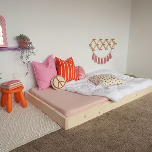 Puede incluir: Una cama baja con una s&aacute;bana bajera rosa claro, un edred&oacute;n blanco y cojines decorativos. Un peque&ntilde;o taburete naranja sostiene libros. Un espejo rosa con bordes ondulados y un perchero de pared de madera adornan la habitaci&oacute;n.