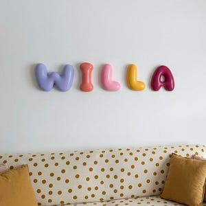 Peut inclure: Un ensemble de cinq lettres murales individuelles formant le nom WILLA. Chaque lettre présente un aspect lisse, arrondi et semblable à un ballon dans des couleurs distinctes : violet clair, corail, rose, jaune et magenta foncé, disposées horizontalement.