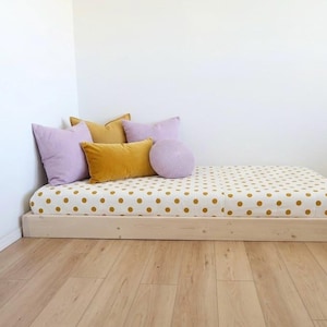 May include: A low-profile wooden platform bed frame featuring a mattress with a white and mustard yellow polka dot pattern. The bed is accessorized with two square lavender pillows, one rectangular mustard yellow pillow, and one round lavender pillow.