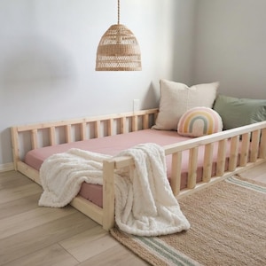 May include: A low-profile wooden floor bed frame with vertical slats, featuring a dusty pink mattress, a white textured throw blanket, a large beige pillow, a sage green pillow, and a small decorative pillow with a rainbow graphic design.