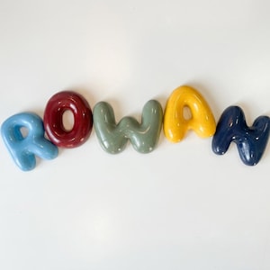 May include: A set of five individual, glossy, rounded, three-dimensional letters spelling the name ROWAN. The letter R is light blue, O is dark red, W is sage green, A is yellow, and N is dark navy blue. Each letter has a smooth, reflective finish.