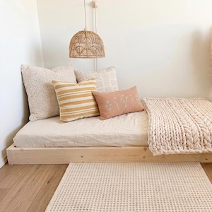 May include: A low-profile wooden platform bed frame with a light-colored mattress, topped with a chunky knit cream blanket, a textured cream pillow, a yellow and white striped pillow, and a small terracotta pillow with white floral embroidery.