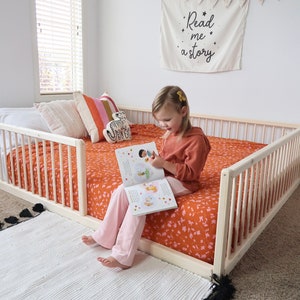 May include: A light wood toddler bed with safety rails. The bed is made up with an orange floral patterned blanket and pillows. A white rug with black tassels is in front of the bed. A banner on the wall reads "Read me a story."
