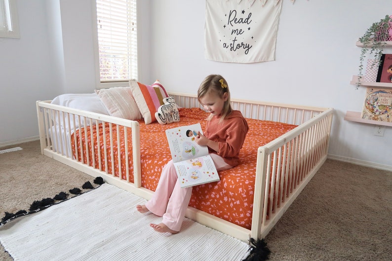 May include: A light wood toddler bed with safety rails. The bed is made up with an orange floral patterned blanket and pillows. A white rug with black tassels is in front of the bed. A banner on the wall reads "Read me a story."