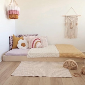May include: A cozy, neutral-toned bedroom setup featuring a low wooden bed frame with pillows and blankets. Decorative elements include a flower-shaped pillow, a rainbow wall hanging, and a wooden rainbow toy. A woven rug and a hanging chandelier complete the space.
