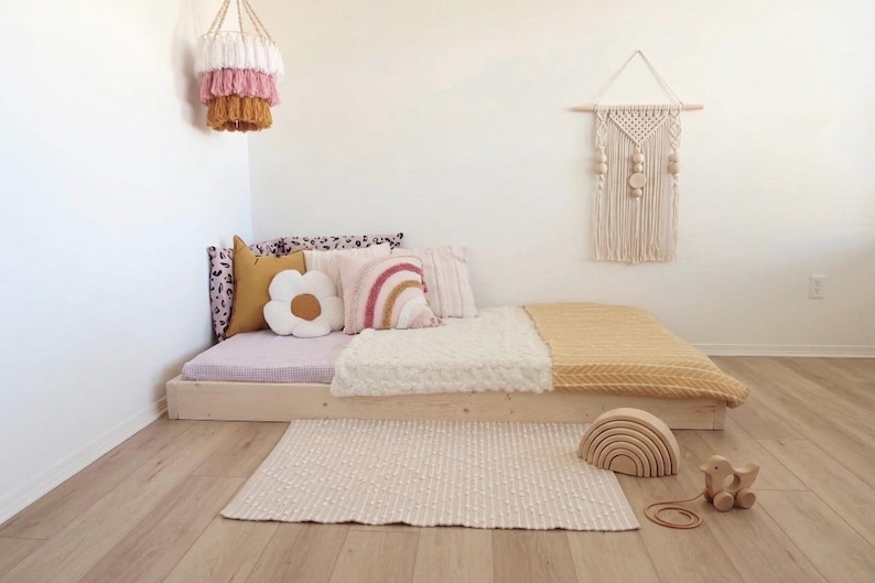 Puede incluir: Un dormitorio infantil de tonos neutros con una cama baja de madera, adornada con cojines y mantas. Elementos decorativos incluyen una almohada en forma de flor, un colgante de pared de arco&iacute;ris y un juguete de arco&iacute;ris de madera. Una alfombra tejida y una l&aacute;mpara colgante completan el espacio.