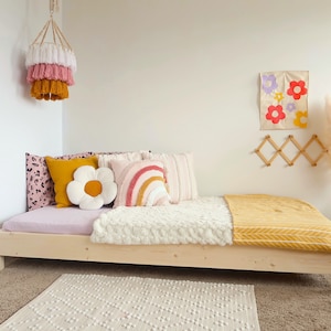 Puede incluir: Una cama de madera clara con una funda de colch&oacute;n estampada en rosa y blanco, adornada con cojines decorativos de varios colores y dise&ntilde;os. Una manta blanca texturizada y una manta amarilla a&ntilde;aden calidez. Una l&aacute;mpara colgante y un arte mural floral completan la decoraci&oacute;n.