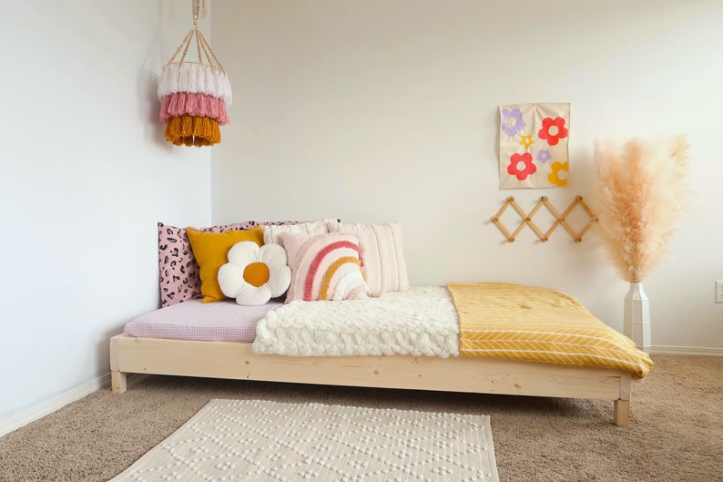 Puede incluir: Una cama de madera clara con una funda de colch&oacute;n estampada en rosa y blanco, adornada con cojines decorativos de varios colores y dise&ntilde;os. Una manta blanca texturizada y una manta amarilla a&ntilde;aden calidez. Una l&aacute;mpara colgante y un arte mural floral completan la decoraci&oacute;n.