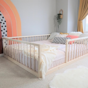 May include: A low-profile, light wood bed frame with vertical slats, surrounded by a matching railing. The bed is made up with a pink sheet, pillows, and a cream-colored blanket. A rainbow mural decorates the wall.