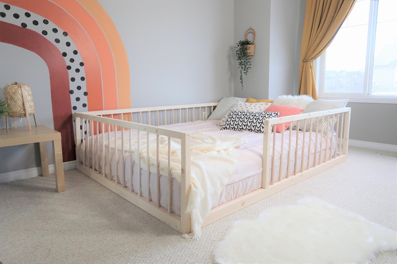 May include: A low-profile, light wood bed frame with vertical slats, surrounded by a matching railing. The bed is made up with a pink sheet, pillows, and a cream-colored blanket. A rainbow mural decorates the wall.