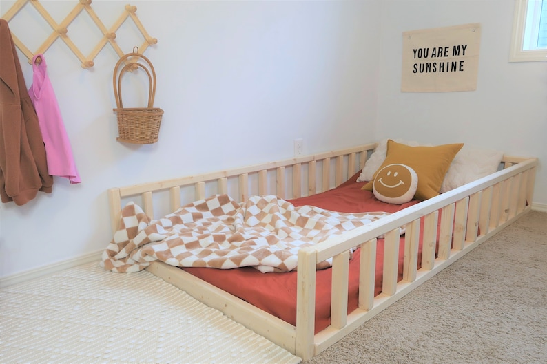Puede incluir: Una cama baja de madera natural con un colch&oacute;n rojo y una manta a cuadros. La cama est&aacute; adornada con una almohada amarilla con una cara sonriente y una almohada color mostaza. Un cartel en la pared dice "YOU ARE MY SUNSHINE".