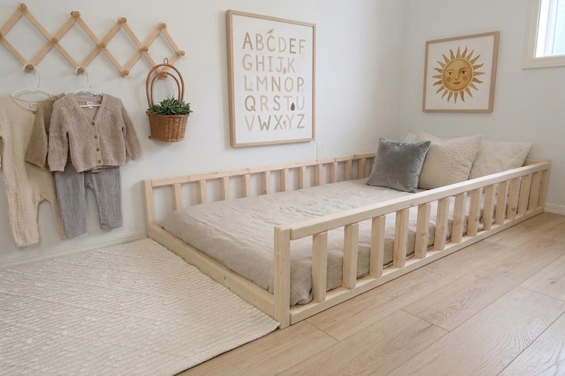 Puede incluir: Una cama con marco de madera clara y perfil bajo, con colch&oacute;n y almohadas beige. La habitaci&oacute;n incluye una alfombra blanca, un perchero de madera montado en la pared y obras de arte enmarcadas. La cama mide unos 200 cm de largo.