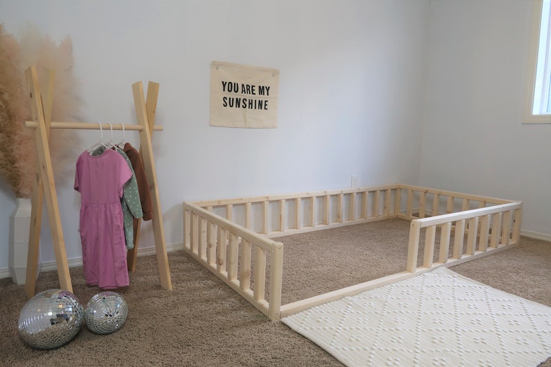 Puede incluir: Un &aacute;rea de juego con marco de madera clara y una alfombra blanca estampada. Un perchero de madera sostiene vestidos en rosa, verde y marr&oacute;n. Dos bolas de discoteca est&aacute;n en el suelo. Una pancarta en la pared dice "YOU ARE MY SUNSHINE".