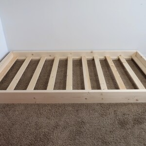 Puede incluir: Un somier de madera sin terminar, de forma rectangular, con un interior de listones. El marco es de madera de color claro y descansa sobre una alfombra marr&oacute;n. El somier est&aacute; contra una pared blanca.