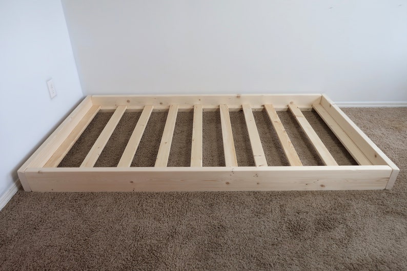 Puede incluir: Un somier de madera sin terminar, de forma rectangular, con un interior de listones. El marco es de madera de color claro y descansa sobre una alfombra marr&oacute;n. El somier est&aacute; contra una pared blanca.
