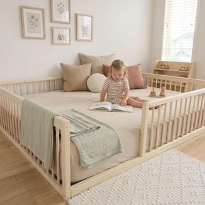 May include: A light wood-framed toddler bed with a mattress, surrounded by a railing. A child is sitting on the bed reading a book. The bed is decorated with pillows and a blanket. The room has a neutral color palette.